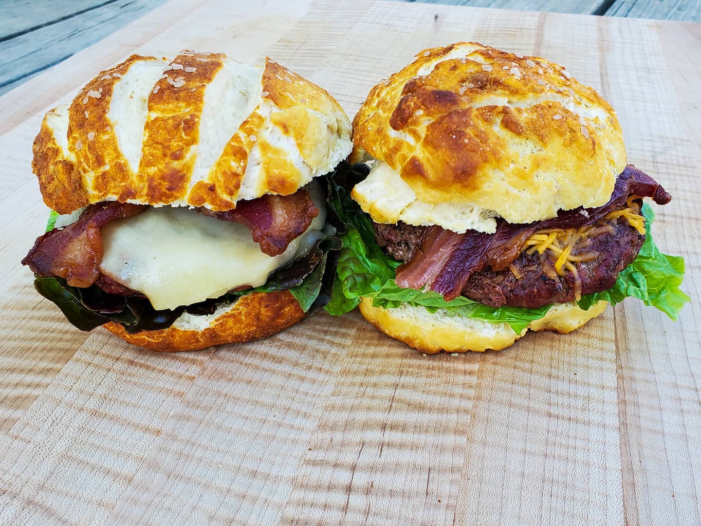 Two burgers with bacon, cheese, and lettuce on a wooden cutting board.