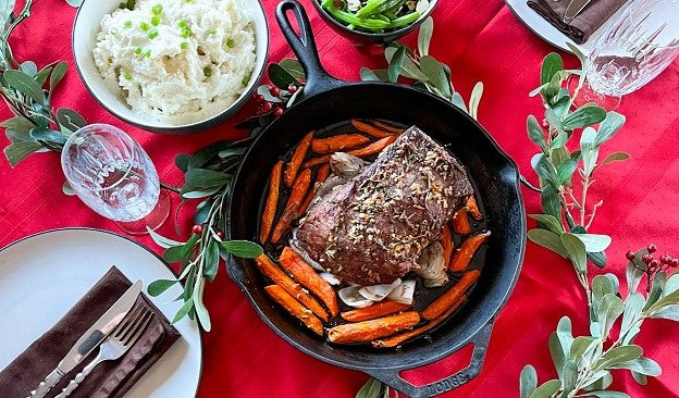 Roast beef with carrots in a skillet on a red tablecloth with side dishes.